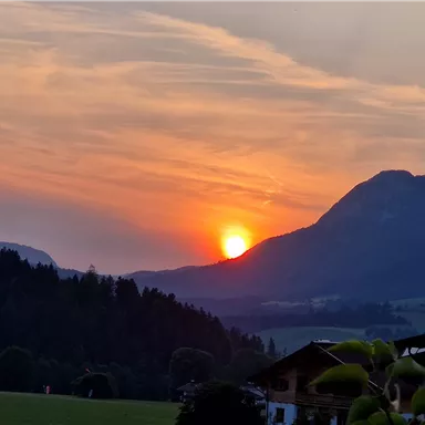 A beautiful sunset behind the mountains, with warm colors in the sky. In the foreground, trees and a building are visible.