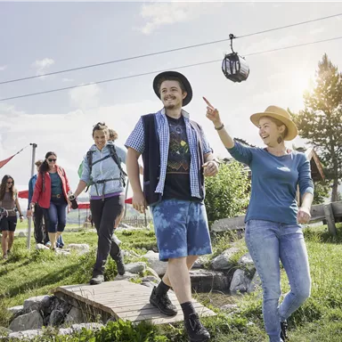 A group of five people is walking on a path in nature. In the background, there is a cable car and a sunny landscape.
