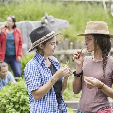 Two women are standing in a garden and chatting amicably. In the background, other people are busy with vegetables.
