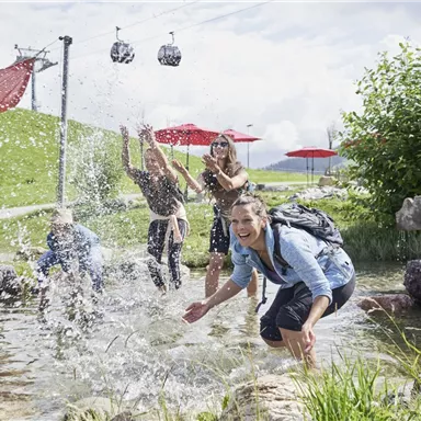 A group of young people is playing in the water and splashing around. In the background, gondolas and red sun umbrellas are visible.
