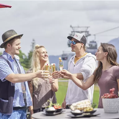 A happy group of four people is toasting with glasses. In the background, there are mountains and a cable car.