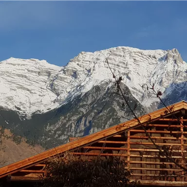 Eine beeindruckende Berglandschaft mit schneebedeckten Gipfeln unter einem blauen Himmel. Im Vordergrund ist ein Holzhaus zu sehen.