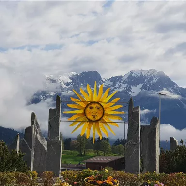 Eine farbenfrohe Sonnenskulptur steht vor majestätischen Bergen, die teilweise von Wolken umhüllt sind. Im Vordergrund sind Blumen zu sehen, die die Szene lebendig machen.