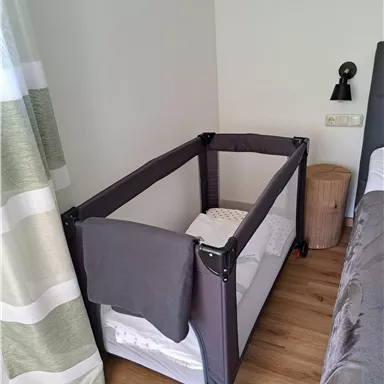 Ein Kinderbett in einem modernen Schlafzimmer. Das Bett ist mit einer Decke ausgestattet und steht neben einem Bett und einem Nachttisch.