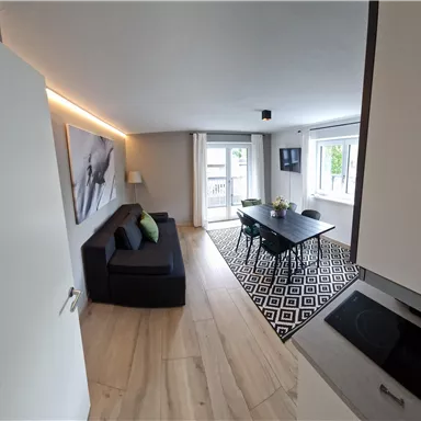Ein modernes Wohnzimmer mit einem Sofa und einem Esstisch. Große Fenster sorgen für viel Tageslicht.
