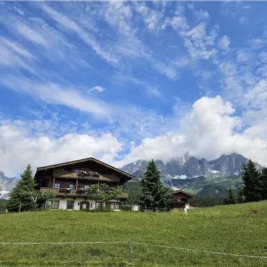 A beautiful farmhouse surrounded by a green meadow and high mountains. The sky is clear with some clouds.