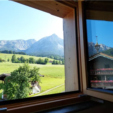 Ein Blick aus einem Fenster auf eine grüne Wiese und majestätische Berge. Im Vordergrund sind ein Holzhaus und Bäume zu sehen.