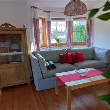 A cozy living room with a comfortable gray sofa and red cushions. A wooden table is in the center, surrounded by large windows that let in plenty of light.