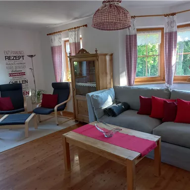 A cozy living room with a light sofa and red cushions. There is a wooden table and windows with curtains that let in plenty of daylight.
