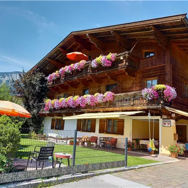 A traditional wooden house with colorful flowers on the balcony. In the foreground, there is a garden with seating areas and sun umbrellas.