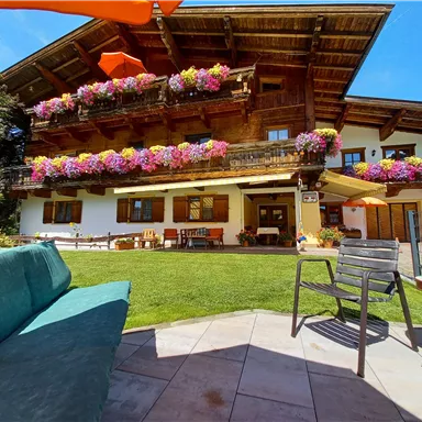 A rustic wooden house with colorful flower boxes and a beautiful garden. In the foreground, there are garden furniture on a terrace.
