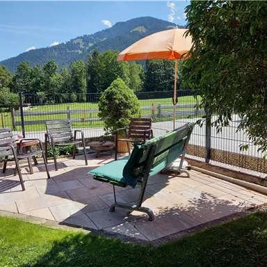 A cozy seating area outdoors with a sun umbrella and several chairs. In the background, green hills and a clear blue sky can be seen.