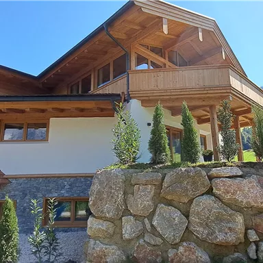 A modern house with wooden cladding and a large balcony. It is situated in a green landscape with mountains in the background.