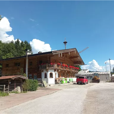 A traditional wooden house with a balcony and red flowers. In the background, trees and a blue sky are visible.