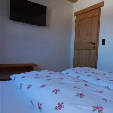 A cozy bedroom with a double bed and floral bedding. A TV is mounted on the wall, and the wooden door leads outside.