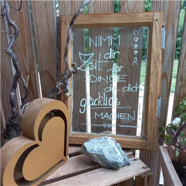 A wooden frame with a handwritten message encouraging you to take time for the things that bring you happiness. Next to it lies a heart-shaped decoration and a stone on a wooden shelf.