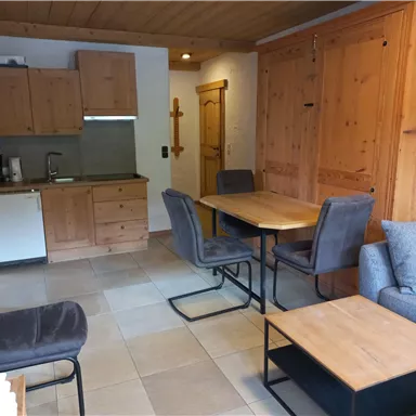 A cozy kitchen with wooden cabinets and a dining table. The living area is furnished with comfortable chairs and a couch.