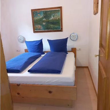 A cozy bedroom with a double bed and blue pillows. A landscape painting hangs on the wall, and the room feels inviting and bright.