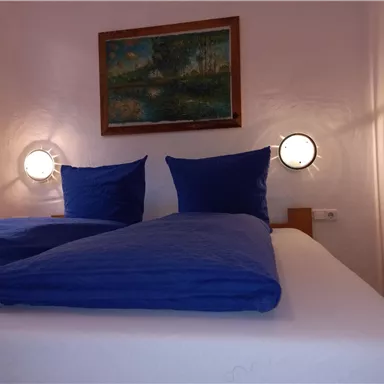 A cozy bedroom with two single beds and blue bed covers. A painting is hanging on the wall and there are two light sources next to the beds.