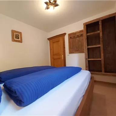 A simple bedroom with a double bed and blue pillows. There is a wooden wardrobe and a warm glow from the ceiling light.