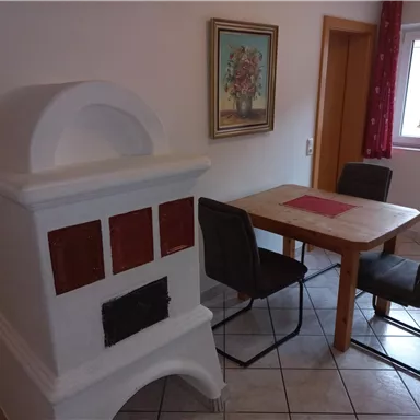A cozy dining area with a wood stove and a table with chairs. A painting hangs on the wall, and the window brings in natural light.