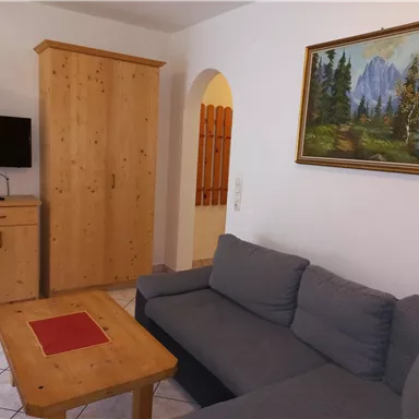 A cozy living area with a gray couch, a wooden table, and a TV. A picture of a mountain landscape hangs on the wall.
