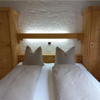 A cozy bedroom with a double bed and wooden cabinets. The pillows have a special shape and there is soft light behind the headboard.