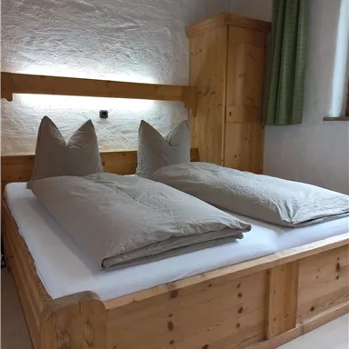 A cozy bedroom with a wooden bed and large, gray pillows. The walls are light and there is a wooden wardrobe as well as a curtain.