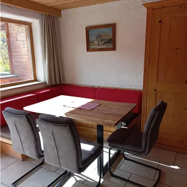 A cozy dining area with a wooden table, surrounded by gray chairs. The walls are white with a red upholstered seating area and a window looking outside.