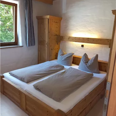 A cozy bedroom with a wooden double bed and gray bedspreads. The room has a window overlooking greenery and warm lighting.