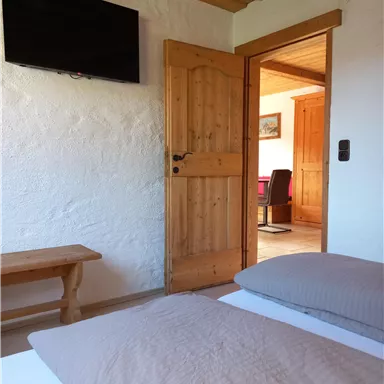 A cozy bedroom with two beds and a wooden chair. Through the open door, you can see another room with a desk.