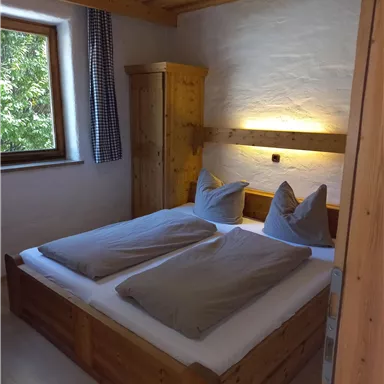 A cozy bedroom with a wooden double bed and large pillows. The room has a window that lets in natural light.
