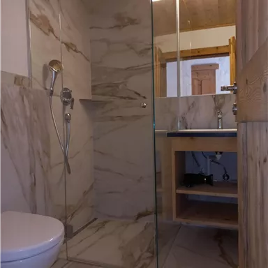 A modern bathroom with a transparent shower and elegant marble elements. There is a toilet and a wooden base with storage space.
