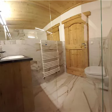 A modern bathroom with marble floors and a glass shower. The walls are light and there is a wooden ceiling as well as a wooden sliding door.