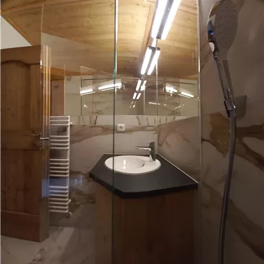 A modern bathroom with a glass door and an elegant sink. The walls are designed with marble and wood, and there is a shower with a rain showerhead.