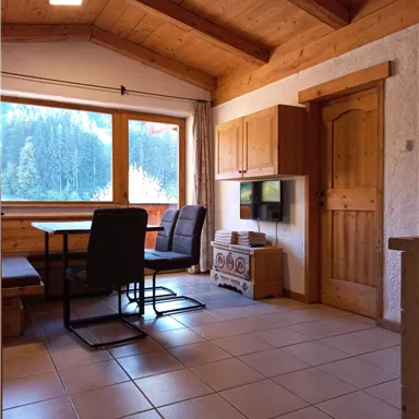 A cozy room with a wooden ceiling and large windows. A table with chairs is in the middle, and there is a television on the wall.