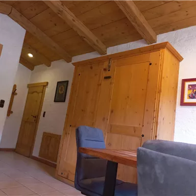 A cozy living space with wood paneling and a large wardrobe. The room includes a table and chairs, as well as a door that leads to another area.