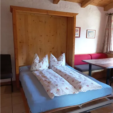 A cozy room with a foldable bed and two pillows. There is wood paneling on the wall, and there is a dining table with chairs.