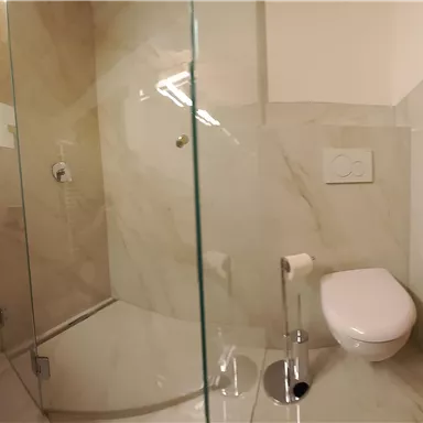 A modern bathroom with a glass shower enclosure and an elegant toilet. The walls are clad with bright, marbled tiles.