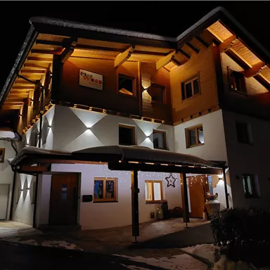 Ein modernes Haus mit Holzdetails, das bei Nacht beleuchtet ist. Der Schnee auf dem Boden und die Lichter verleihen der Szene eine gemütliche Atmosphäre.