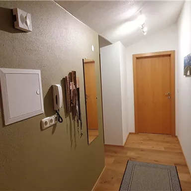 Ein schmaler Flur mit Holzboden und einer grünen Wand. Mehrere Türen führen in verschiedene Räume, und an der Wand hängen einige Gegenstände.