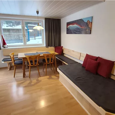 A cozy living space with a wooden table and chairs. A sofa with red cushions and large windows provide natural lighting.