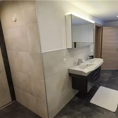 A modern bathroom with a shower and a large mirror. The walls are in a light stone look and the floor is dark.