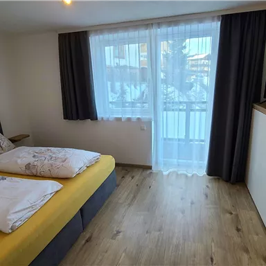 A modern bedroom with two beds, a television, and a balcony. Large windows provide plenty of daylight.
