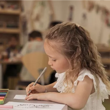 A girl with curly hair is sitting at a table and drawing. In the background, other children and crafting materials are visible.