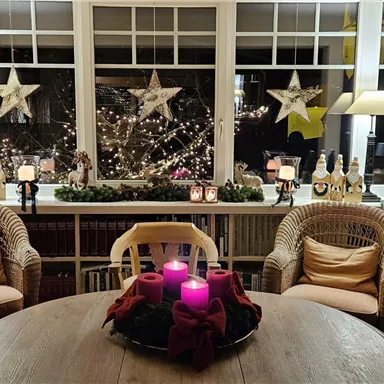 A cozy room with a round table and two basket chairs. The view from the window shows festive decorations, including glowing stars and candles.