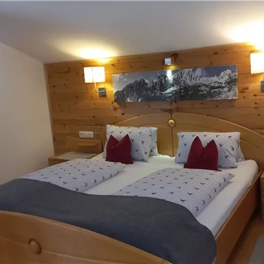 A cozy bedroom with wooden walls and a double bed. The cushions are decorative, and there are nightstands as well as soft lighting.
