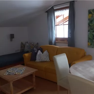 A modern living room with a yellow sofa and a round dining table. There is a sleeping area and a large window with curtains.