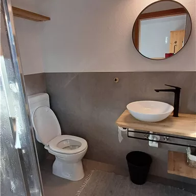 Ein modernes Badezimmer mit einer Dusche, einem Waschbecken und einer Toilette. Die Wände sind in neutralen Tönen gehalten und es gibt einen runden Spiegel.