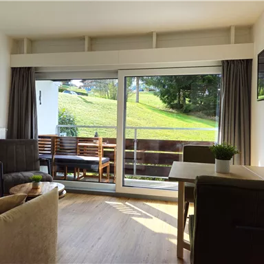 A modern living room with large windows overlooking a green meadow. The room is cozy with a table, chairs, and a TV corner.